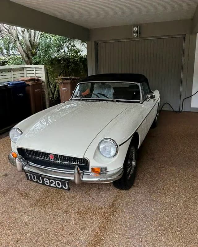 Our white MGB Roadster with its black hood up, parked under a carport